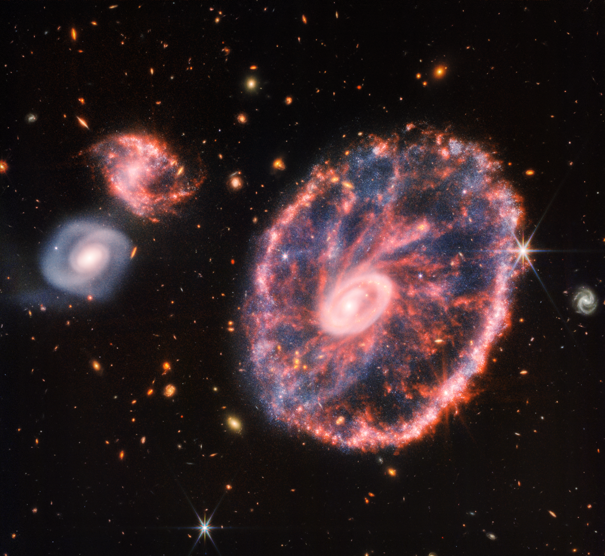 A large pink, speckled galaxy resembling a wheel with a small, inner oval, with dusty blue in between on the right. Two smaller spiral galaxies about the same size are to the left, all against a black background.