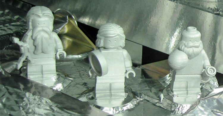 A trio of light gray LEGO figurines stand on platform of silvery metal that extends up behind them. Two bearded men flank a woman in the middle; from left to right they carry a lightning bolt, a magnifying glass, and a telescope and globe of planet Jupiter.