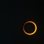 The orange glow of the sun emanates from behind a silhouette of the moon against a dark sky.