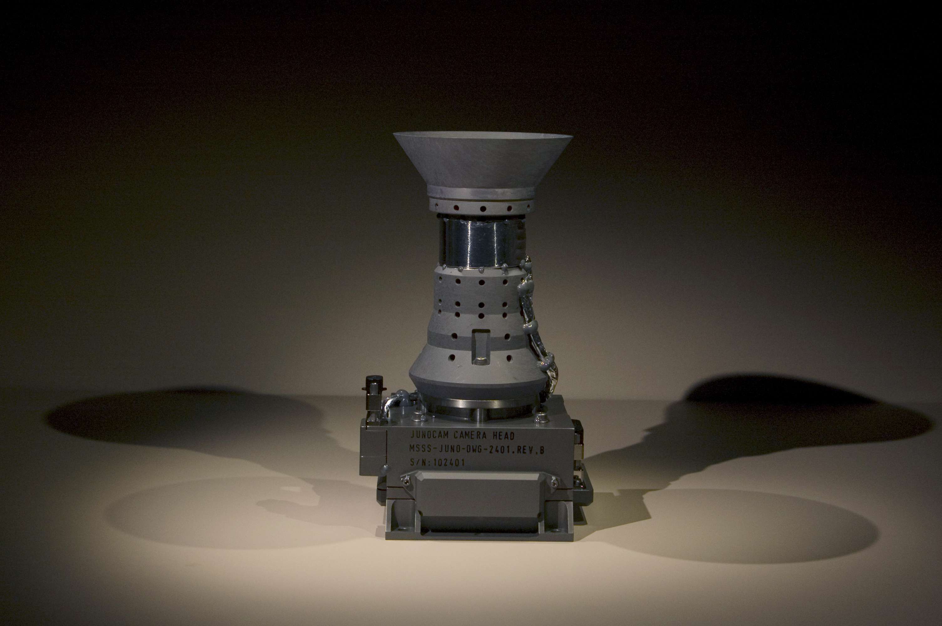 The silver-colored JunoCam camera head sits on a tan table. The cameras lens points up. The camera is sitting on a base inscribed with the name of the camera and some other letters and numbers.