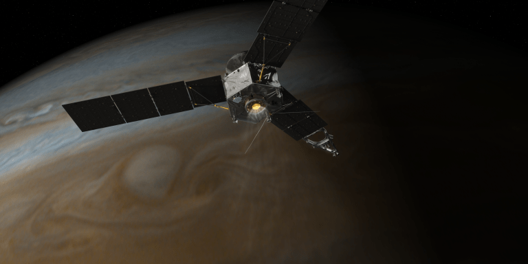 A spacecraft with extended solar panels is orbiting above Jupiter, showcasing the planet’s swirling brown, beige, and white cloud patterns beneath. Jupiter's Great Red Spot is faintly visible, highlighting the spacecraft’s proximity to the gas giant.