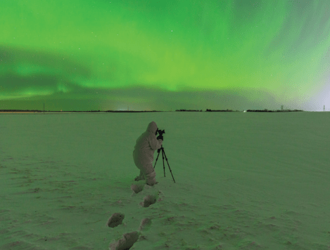 A man in a head-to-toe white snow suit crouches behind a camera on a tripod. He stands knee-deep in snow. The sky above him is filled with a green aurora, which is reflected in the snow.