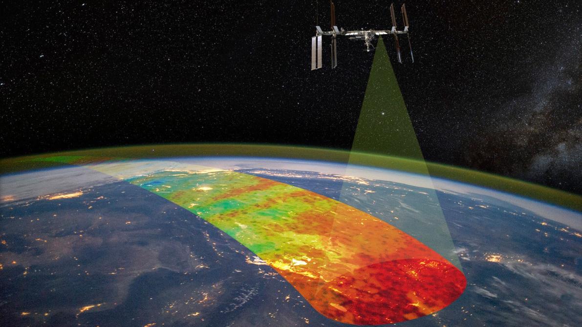 An artist's conception of the international space station flying over earth's atmosphere. A yellowish beam comes down from the space station toward's Earth's atmosphere, leaving a multi-colored path across the sky below it, representing AWE's measurements.