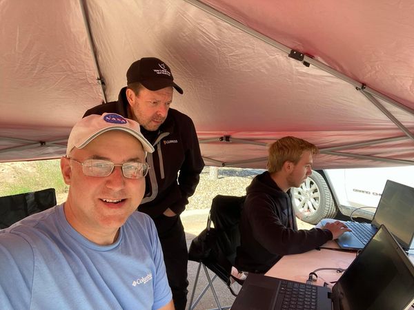 Three men are under an open-sided silvery tent. One is seated, working on a laptop computer on a table. The second is standing, looking down at something on the table. The third, the photographer, smiles happily at the camera, a baseball hat with the round blue NASA logo perched on his head.