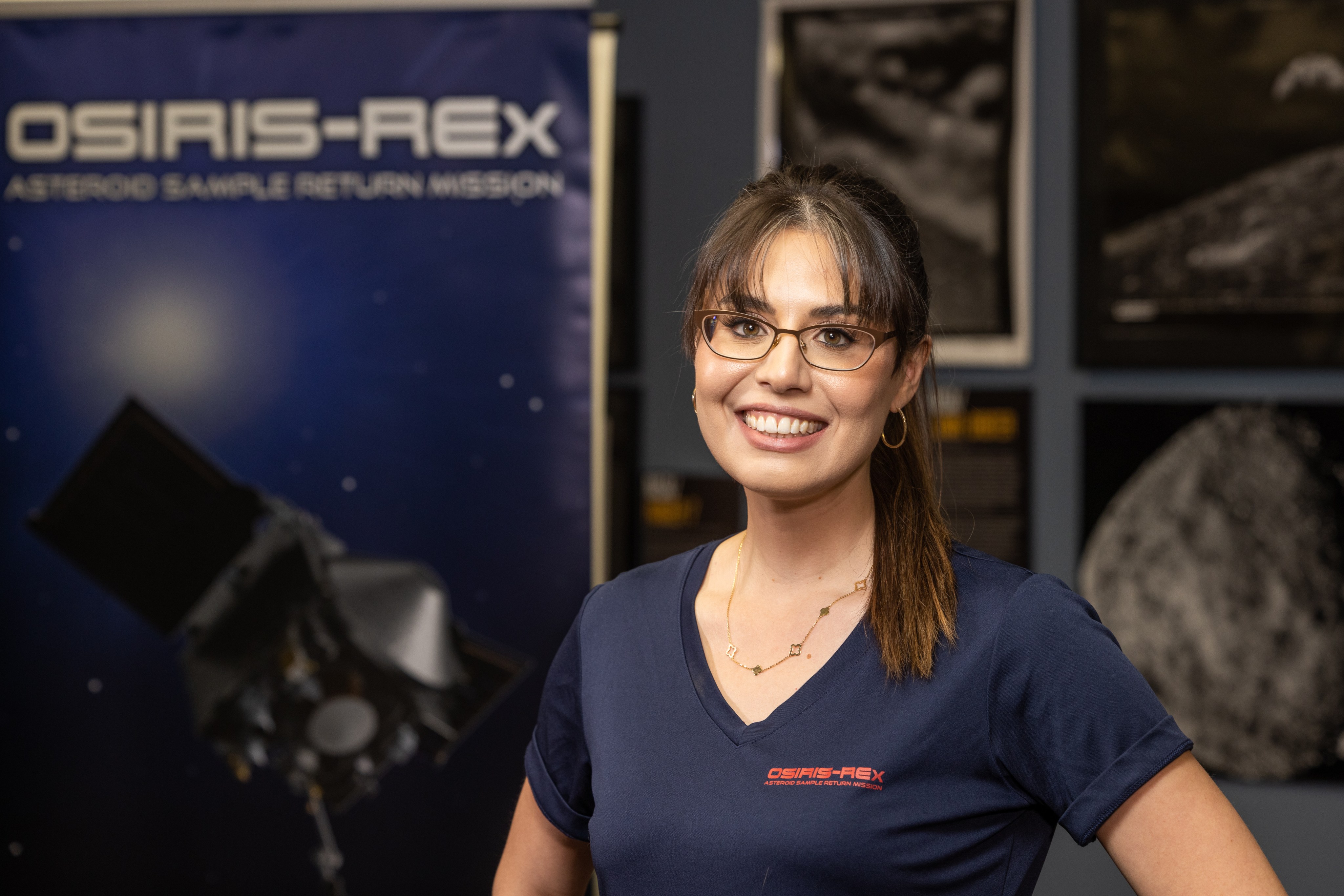 Woman stands in front of OSIRIS-REx poster, wearing a navy blue shirt with OSIRIS-REx in red. She has brown hair and is wearing glasses.