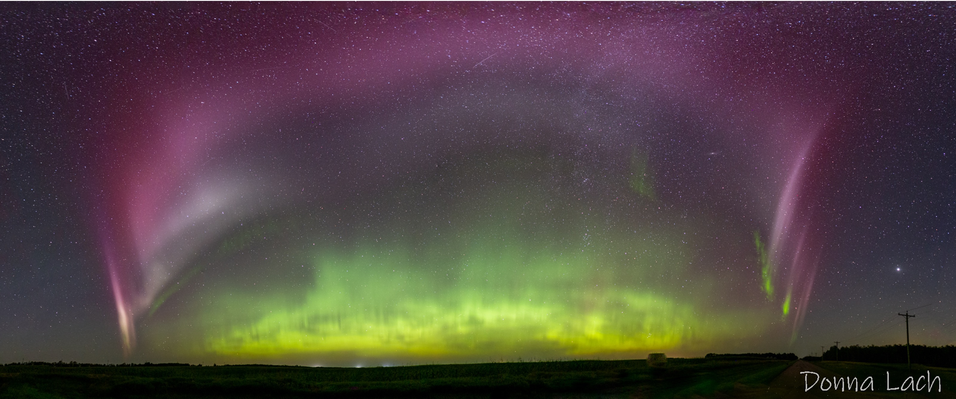 In a starry night-time panorama, a purple and grey arc stretches from side to side (east to west) over a silhouetted plain, framing green aurora in the distance. Directly beneath the arc are green stripes, the “picket fence” features that occur with STEVE.
