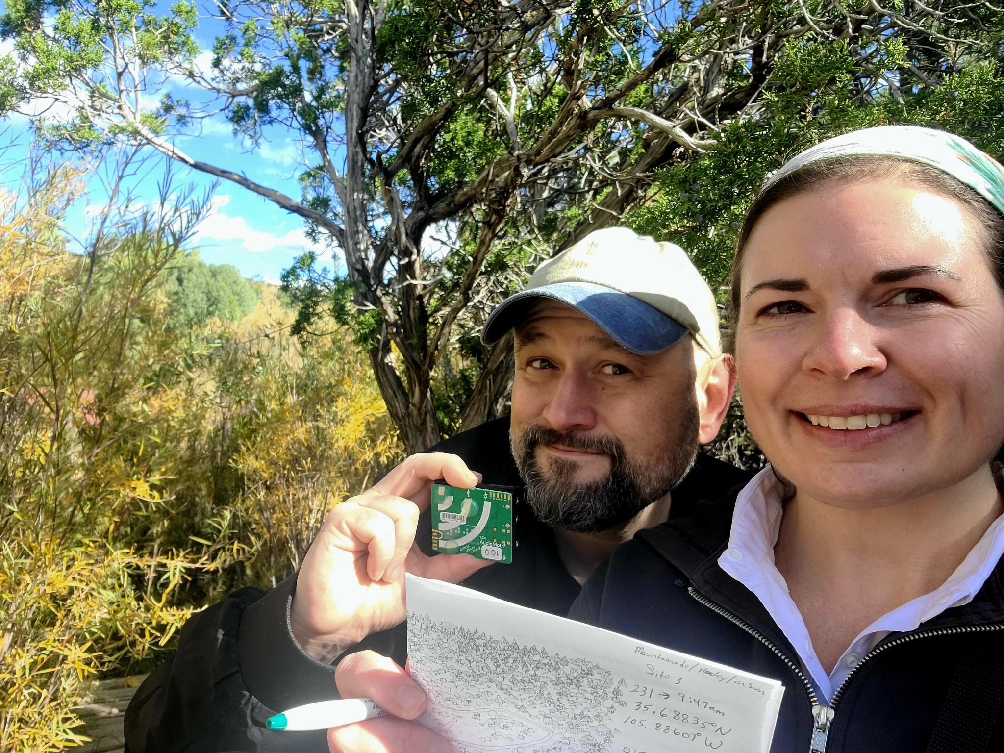 A bearded man in a baseball hat smiles at the camera while he holds up an AudioMoth recording device, which is a small rectangular device about half the size of a cell phone. Next to him, a woman holding a pen and paper covered in notes. She’s also looking at the camera and smiling. The two are under an evergreen trees and surrounded by shrubs with little, yellowing leaves.