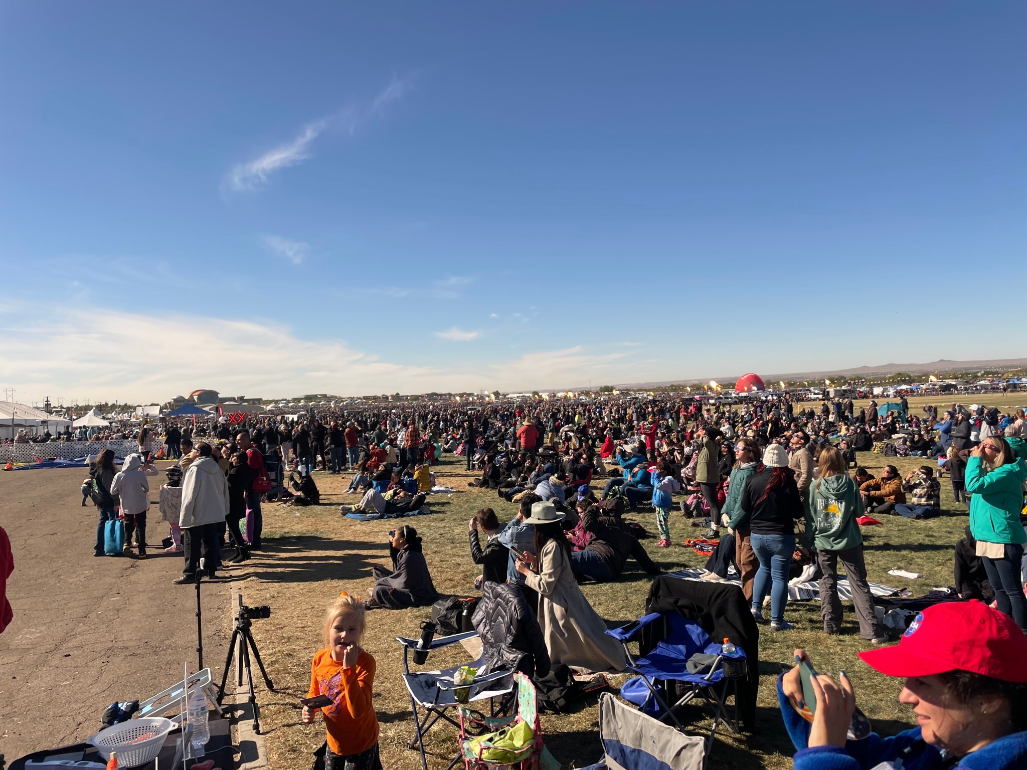 On a dry and dusty open space, a huge crowd of people has gathered, both standing and sitting, many looking up at the sky. The sky above is blue, with some wispy clouds down by the horizon. On the far left some white tent roofs are visible. In the distance on the right we can see a partially inflated red hot air balloon, resting on the ground.