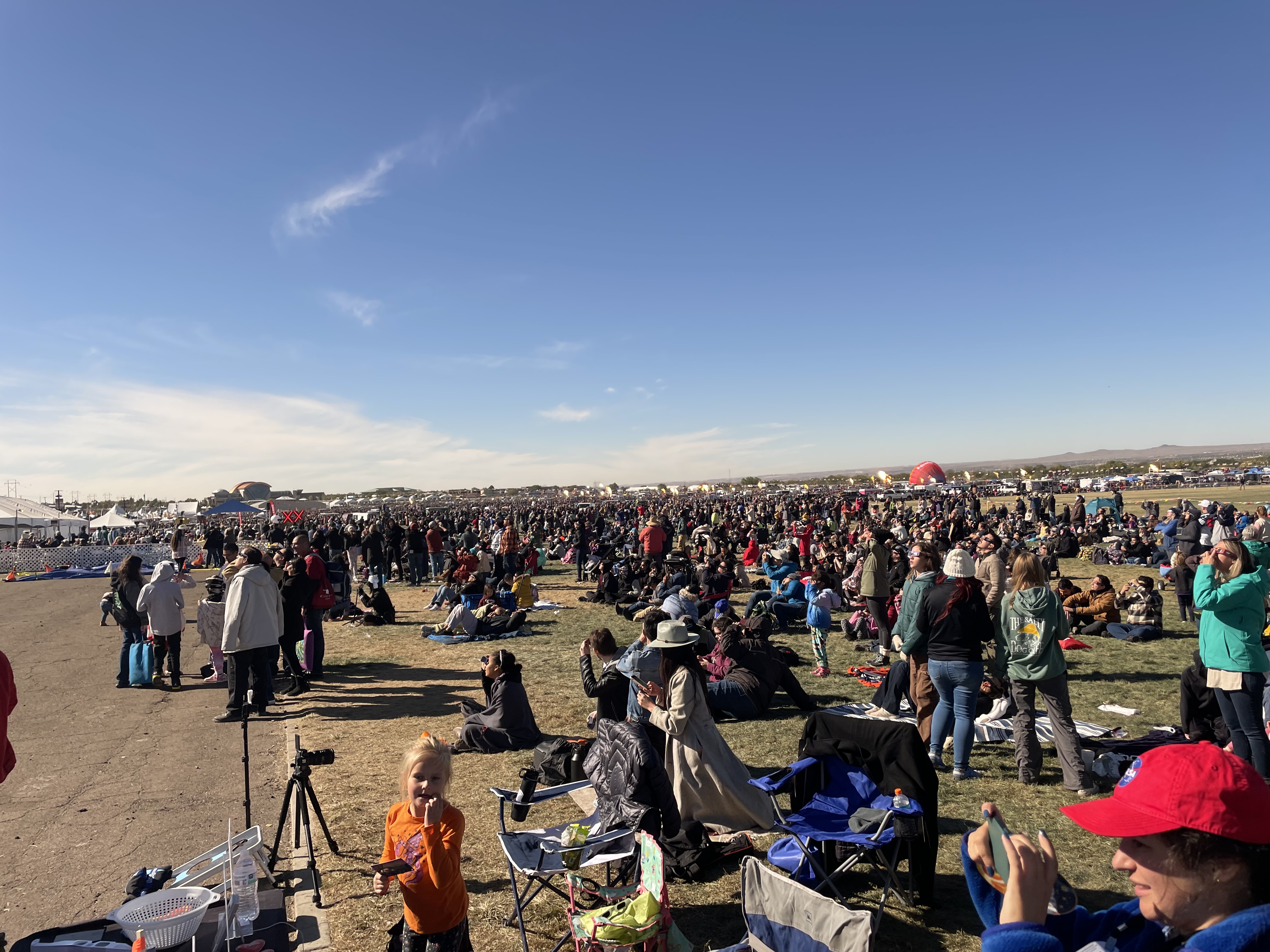 On a dry and dusty open space, a huge crowd of people has gathered, both standing and sitting, many looking up at the sky. The sky above is blue, with some wispy clouds down by the horizon. On the far left some white tent roofs are visible. In the distance on the right we can see a partially inflated red hot air balloon, resting on the ground.