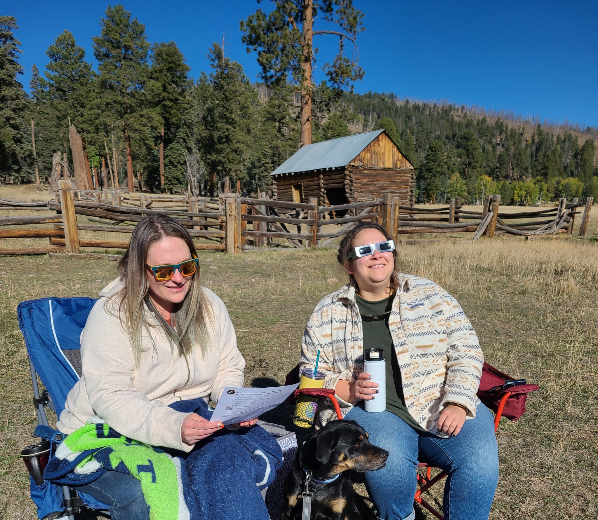 Two women sit on folding camp chairs outside. Behind them is a wooden fence and a small barn built of logs. The grass in the field is golden rather than green. The woman on the left has straight blonde hair and she’s looking at a paper in her hands. The woman on the right, who has brown hair and is wearing eclipse glasses, is looking up at the sky. Both are smiling. A black and brown dog sits on the ground between the two women.