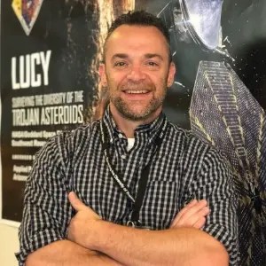 man wearing a black and white checkered button down smiles in front of Lucy poster