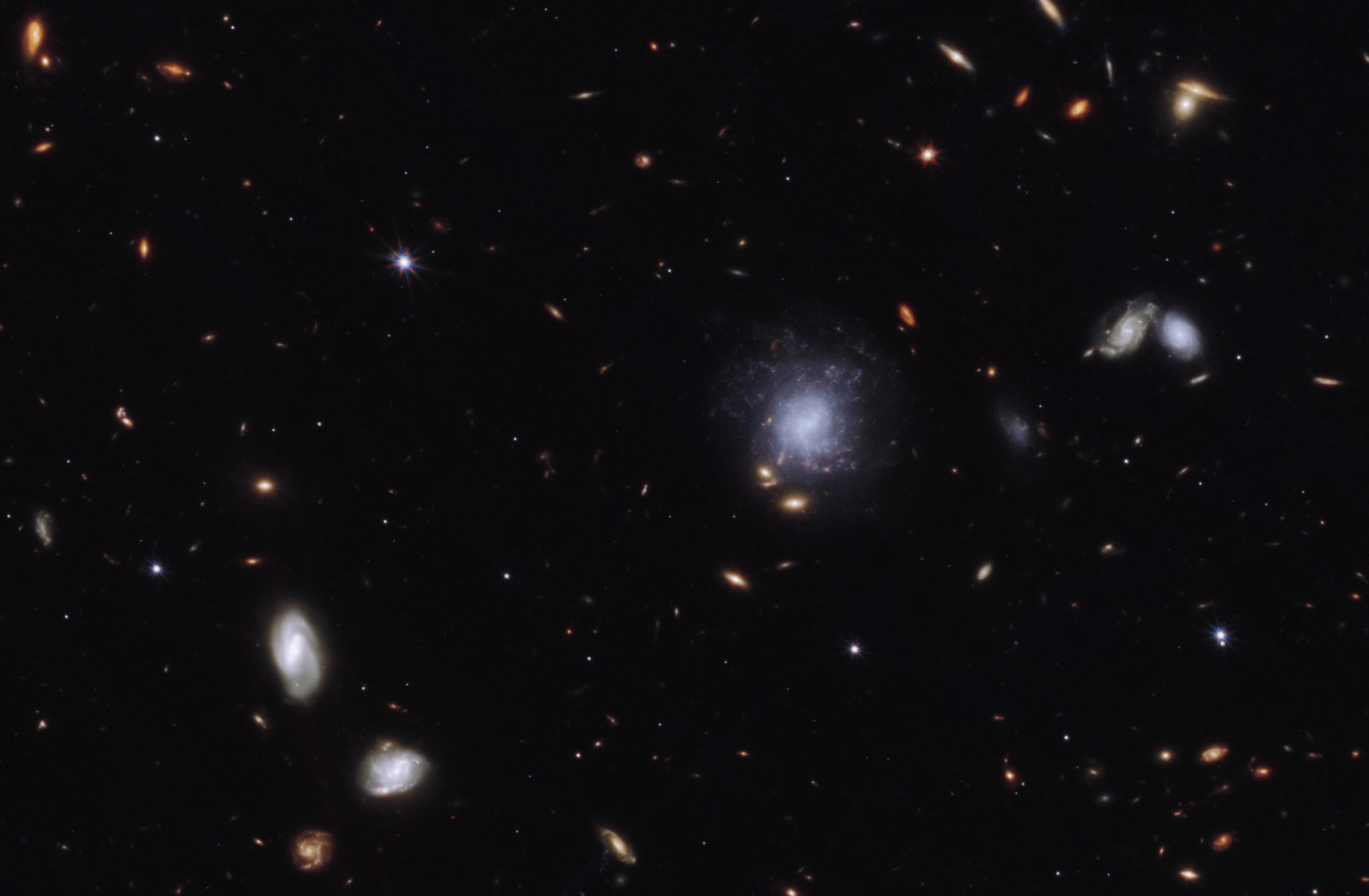 Bright galaxies and other light sources in various sizes and shapes are scattered across a black swath of space: small points, hazy elliptical-like smudges with halos, and spiral-shaped blobs. The objects vary in color: white, blue-white, yellow-white, and orange-red. Toward the center right is a blue-white spiral galaxy seen face-on that is larger than the other light sources in the image. The galaxy is labeled “former home galaxy.” Toward the upper left is a small red point, which has a white circle around it and is labeled “GRB 230307A kilonova.”