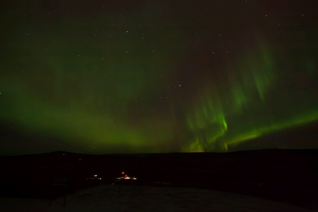 A time-lapse of auroral activity looks like green, red, yellow, and orange curtains traveling across a clear night sky filled with stars. In the dark land below, faraway launch pads are lit by floodlights, and two tiny vertical rockets can be seen waiting for launch.