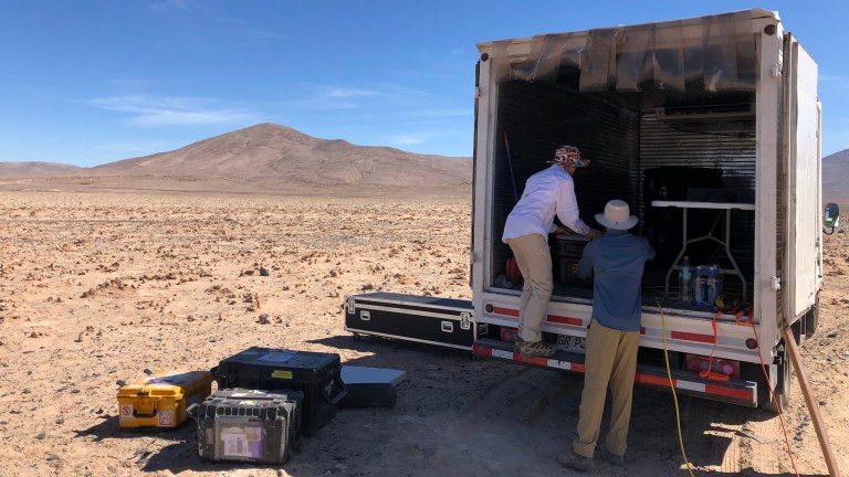 Two scientists unload gear from the back of a truck and onto the desert floor.