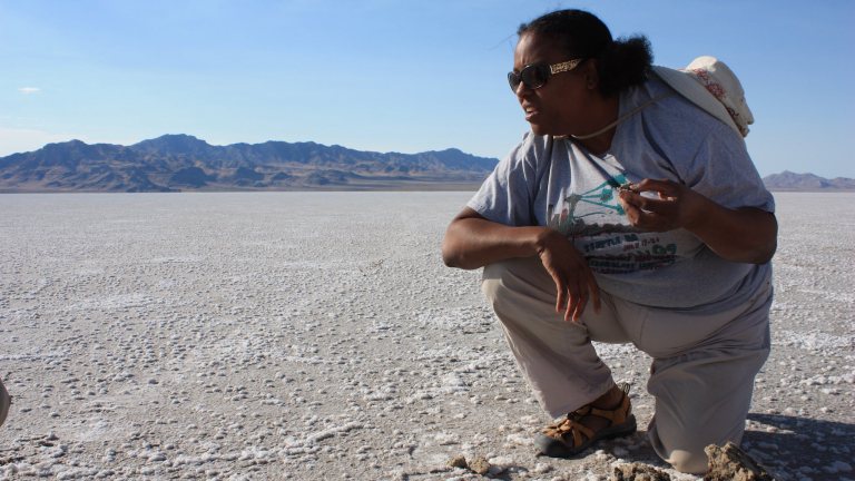 A scientist in a t-shirt and long pants with a hat hanging off her neck inspects rocks in the desert.