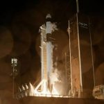 At night, a large, white rocket stands straight up in the middle of the image. On either side are tall towers. The bottom of the rocket is bright, as if it's lighting up, and golden smoke starts to fill the bottom of the image.
