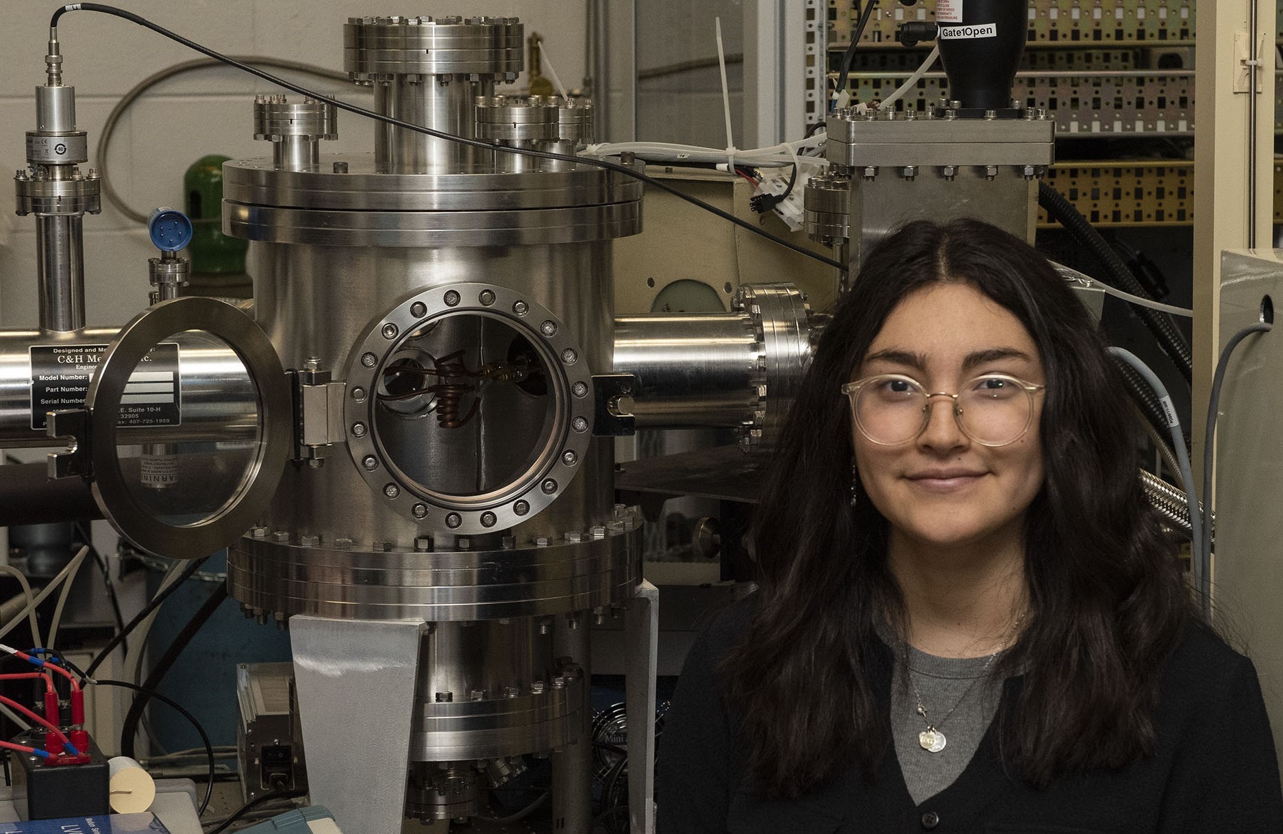 A woman with dark hair and glasses stands in front of a machine in what looks to be a lab.