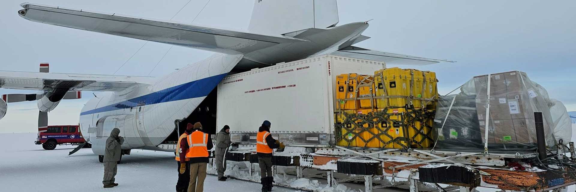 A white airplane sits on the snow at McMurdo Station, Antarctica. Workers, some wearing orange vests, stand near a big white cargo container which is in the tail of the plane. Several smaller yellow and orange containers are behind the white container. They are wrapped in netting. Some brown boxes under plastic are strapped down behind the other containers.