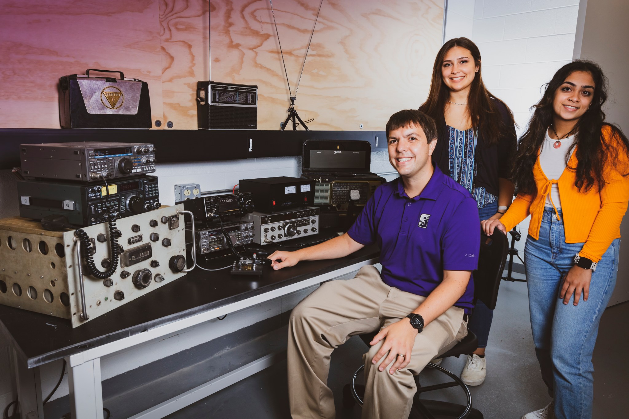 A man in a purple shirt sits at a black-topped table covered in stacked metal boxes covered in knobs and switches. Behind him stand two young women. All three are smiling. 