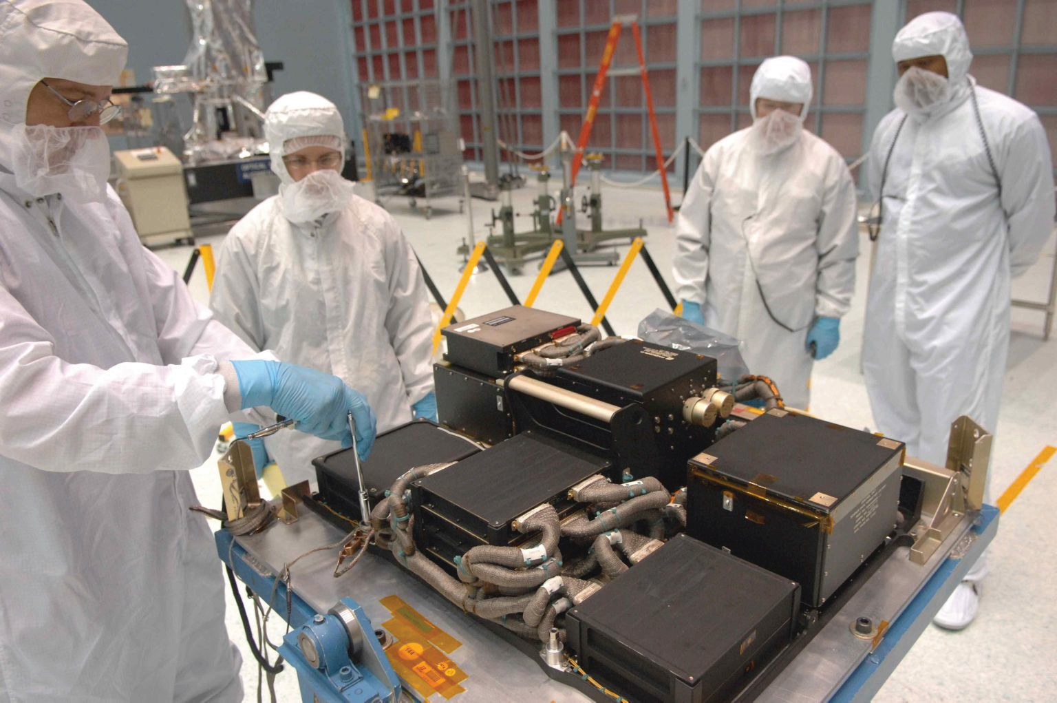 Four people in white suits and masks are in the Goddard Space Flight Center clean room. They are standing around a table that holds a large instrument package, Hubble's Science Instrument Command and Data Handling Module. The module holds several black boxes of varying sizes with bundled wires in tubes connecting them.