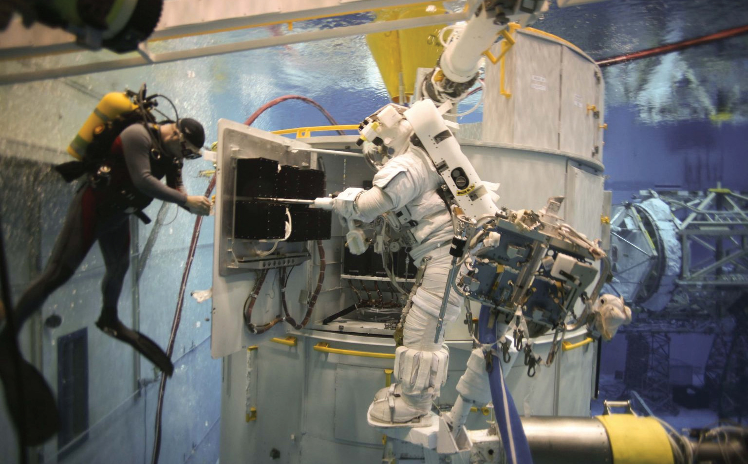 A mock-up of the Hubble Space Telescope in an enormous tank of water. An astronaut, standing on a replica of the Space Shuttle's robotic Canadarm is practicing the installation of Hubble's Science Instrument Command and Data Handling Module. On the far left, a scuba diver works on another component.