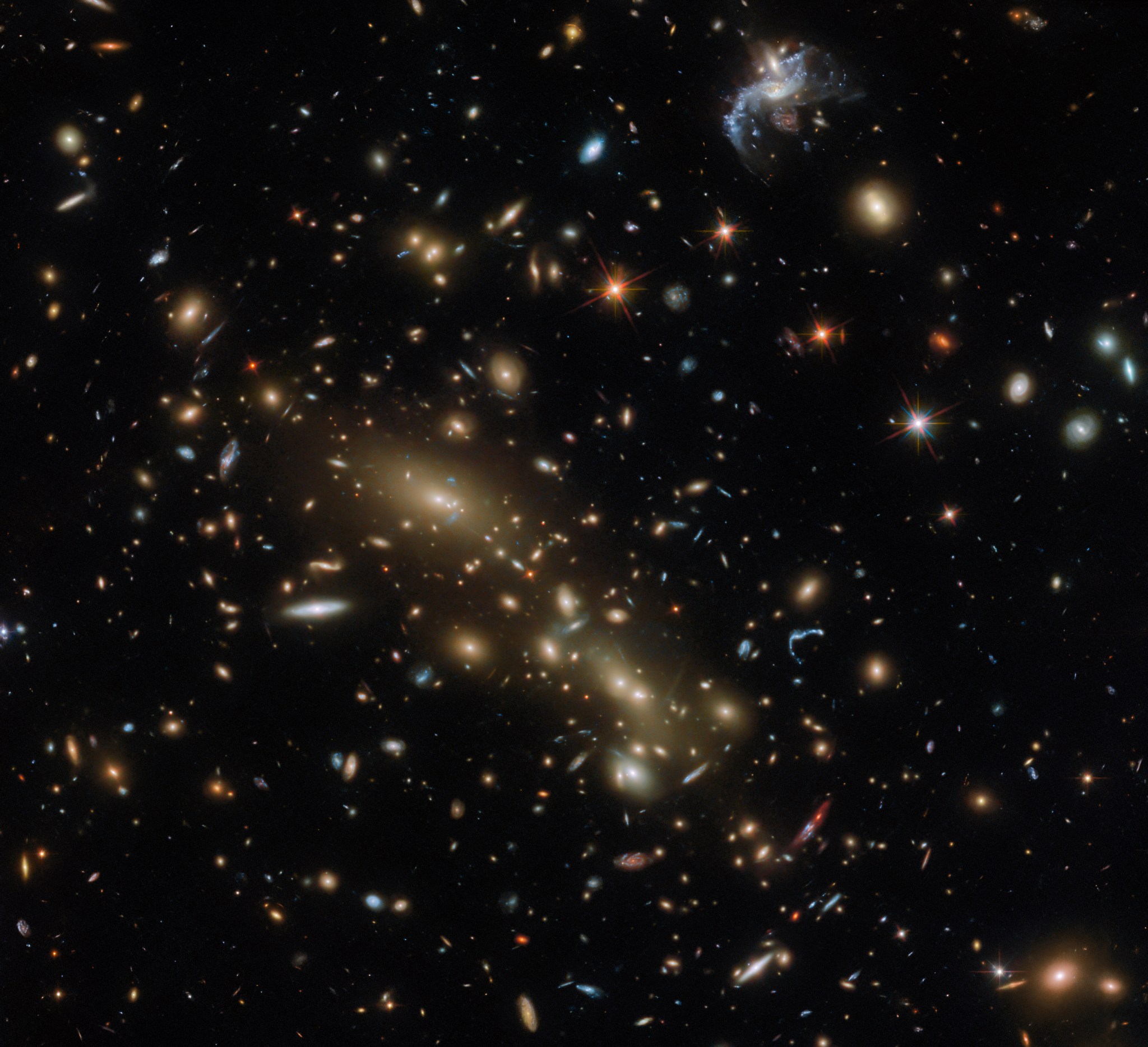 A cluster of galaxies, concentrated around what appear to be two large elliptical galaxies. The rest of the black background is covered in smaller galaxies of all shapes and sizes. In the top left and bottom right, beside the two large galaxies, some galaxies appear notably distorted into curves by gravity.