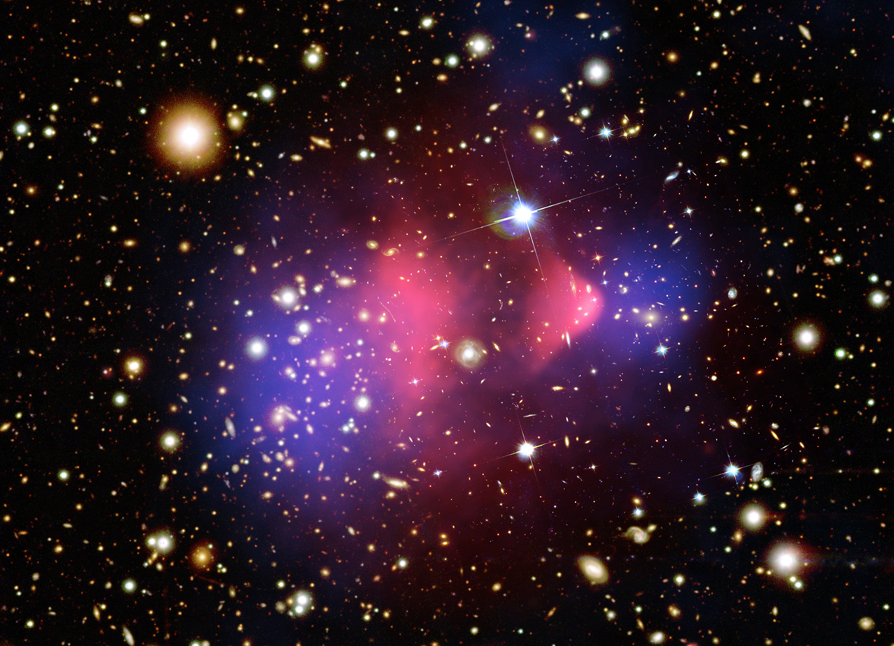 Galaxies fill the scene against a black background. An oval, translucent blob of bright-pink at image center that is edged with purple blobs on either side of the pink.