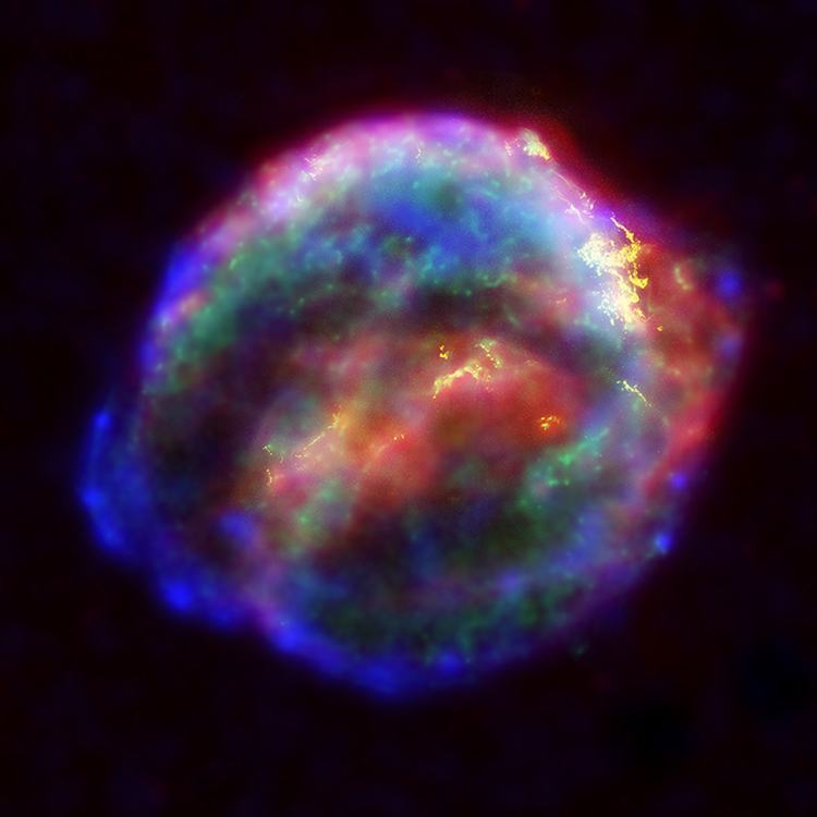 A spheroidal cloud. Its center holds colors of red and yellow with small areas of green and blue. Around the center is a ring of blue and green with a smattering of pink, purple, and white.