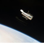 Hubble, at top center, against the black background of space. Earth limb is visible in the lower-left corner.