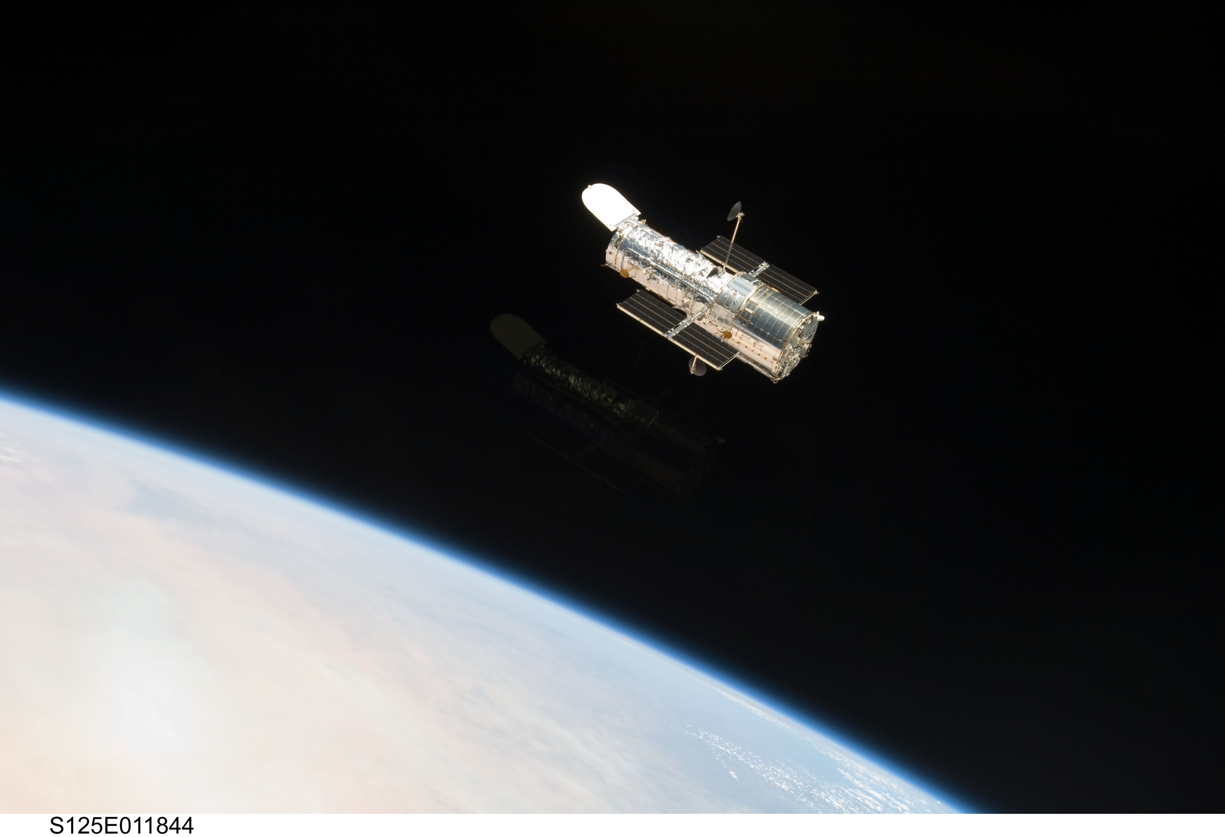 Hubble, at top center, against the black background of space. Earth limb is visible in the lower-left corner.