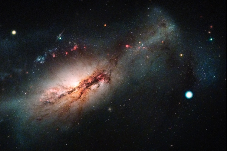A large galaxy fills the frame from the lower-left to the upper right. It appears as bright, pinkish spherical cloud bisected by dark, reddish brown dust lanes and dotted with areas of bright-pink. Below and to the right is a bright-white circle/dot.