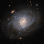 A spiral galaxy, seen face-on from Earth. The spiral arms of the galaxy are bright but not well defined, merging into a swirling disk with a faint halo of dimmer gas around it. The core glows brightly in a lighter color and has a bit of faint dust crossing it. Two redder, visually smaller galaxies and a bright star are prominent around the galaxy, with more tiny objects in the background.