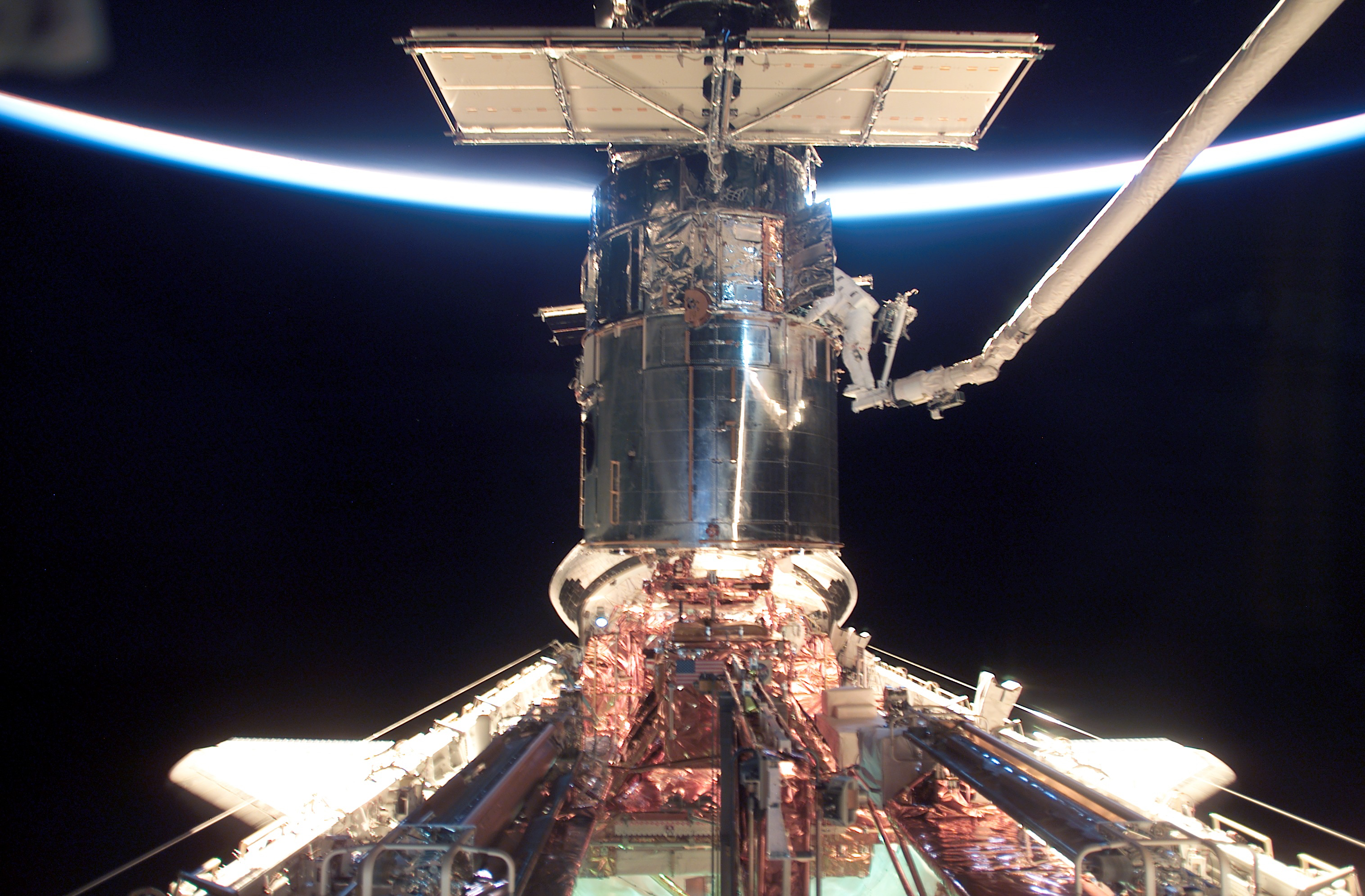 The brightly lit limb of the Earth crosses the top of the image, while the rest of the Earth is dark. The space shuttle is at the bottom of the image with the cargo bay open. Hubble is perpendicular to the shuttle. An astronaut is on the robotic CANADARM to the right of Hubble.