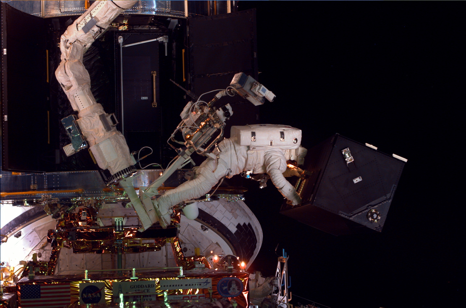 An astronaut on the end of the space shuttle's robotic CANADARM above the shuttle's cargo bay. He is holding Hubble's Faint Object Spectrograph, which looks like a black Hubble is in the background.