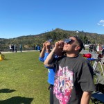 Two people stand looking up at the Sun, while holding solar filters against their eyes.