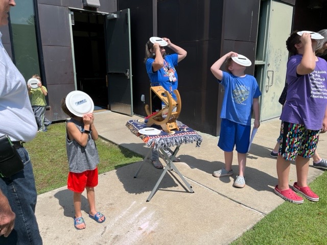 Four children stand with paper plates covering their faces. They are looking up toward the Sun.