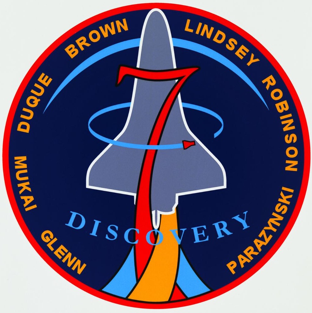 A circular patch with a blue background and a red edge. Along the outer edge of the patch are the names of the crew. Through the center of the patch is a light, bluish-grey line drawing of the space shuttle launching.