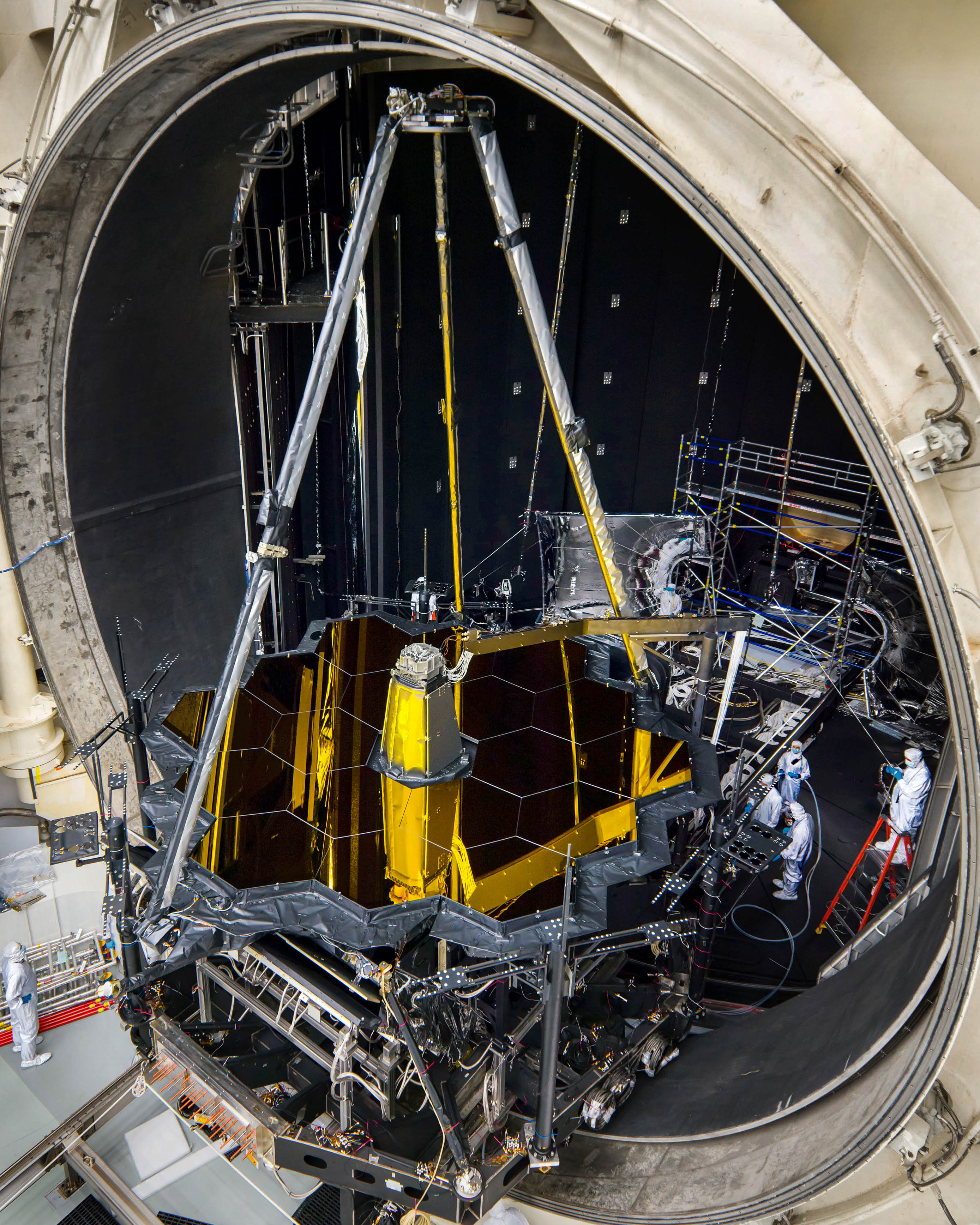 The Webb telescope enters the giant Chamber A thermal vacuum chamber at NASA Johnson. At this point, Webb consists of mirrors and instruments but has not yet been mated with the sunshield or spacecraft bus. Webb is on its back, golden hexagonal mirrors face up. The secondary mirror support structure is extended like a tripod above the primary mirrors. The telescope lies on black and silver support equipment. It is half in the giant mouth of the cavernous test chamber. The chamber is filled with test equipment and people in cleanroom suits. One of them stands on top of a red ladder.