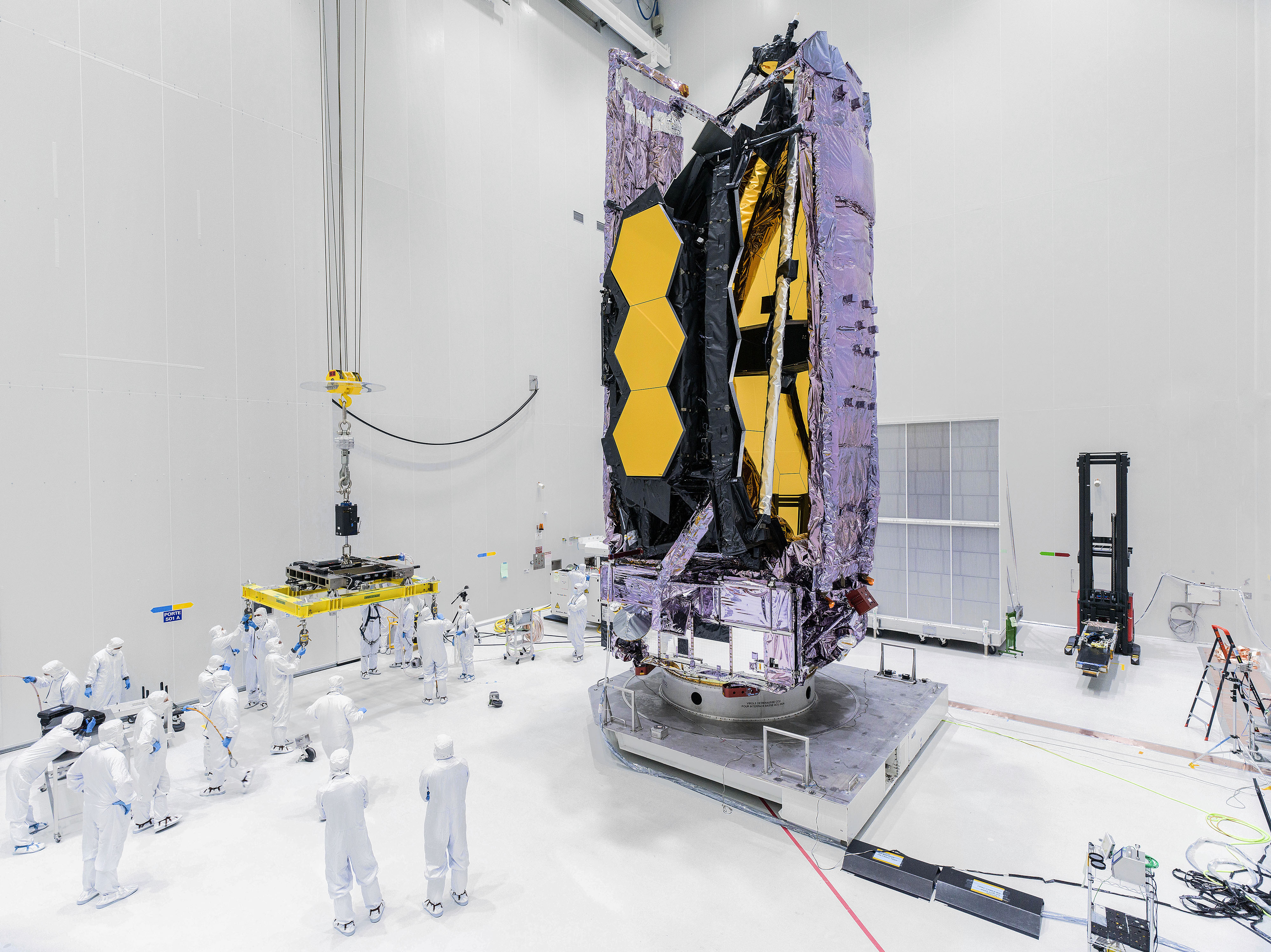 Image taken in a cleanroom at Europe’s Spaceport in Kourou, French Guiana. Dozens of cleanroom technicians, wearing white contamination-control suits and blue gloves, stand towards the left side of this image. On the right side is the Webb telescope in its folded configuration. Its purple sunshield pallets enclose its hexagonal, gold-colored mirror segments. Other hardware, wires, and equipment are located around the cleanroom, which features bright white walls.