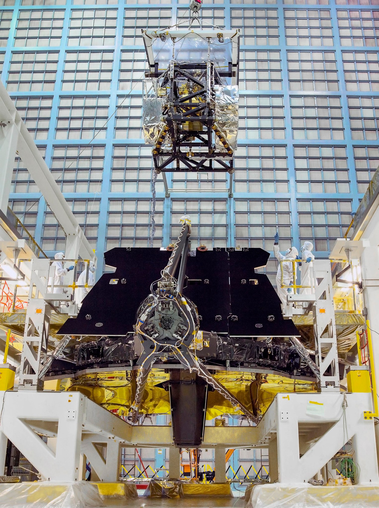 Webb’s instrument module is integrated onto the back of the telescope, right behind the mirrors. The telescope is facedown on a white stand, with the golden mirrors facing down. The secondary mirror support structure is folded over the mirrors, with the round back of the secondary mirrors facing the camera. Behind it is a black radiator panel. The instrument module is being lowered by a crane. The 4 science instruments are visible through the module structure. In the background is a wall of HEPA filters that help keep the NASA Goddard cleanroom clean.