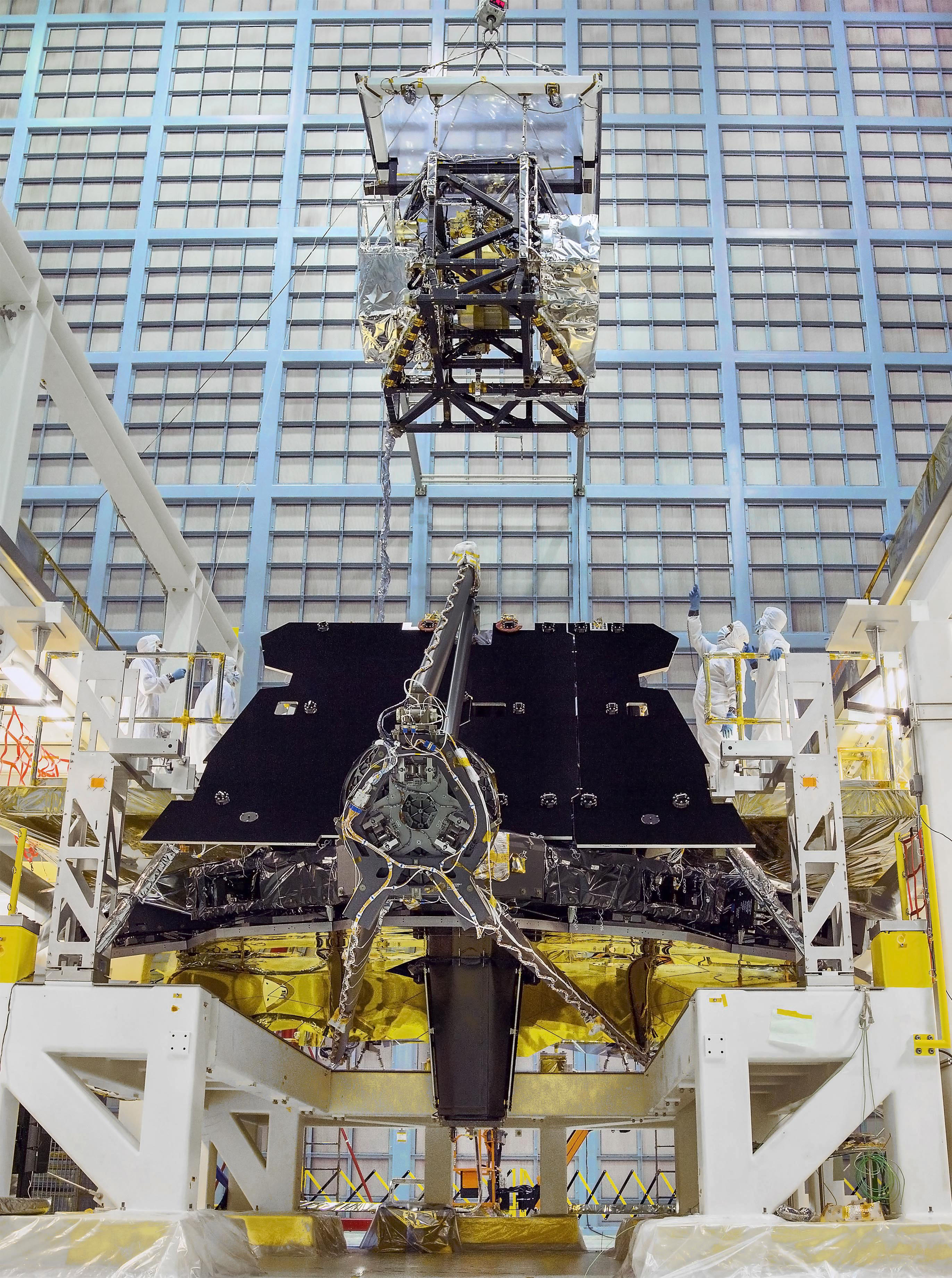 Webb’s instrument module is integrated onto the back of the telescope, right behind the mirrors. The telescope is facedown on a white stand, with the golden mirrors facing down. The secondary mirror support structure is folded over the mirrors, with the round back of the secondary mirrors facing the camera. Behind it is a black radiator panel. The instrument module is being lowered by a crane. The 4 science instruments are visible through the module structure. In the background is a wall of HEPA filters that help keep the NASA Goddard cleanroom clean.