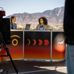 Two women sit at a desk outside. The desk is decorated with art that represents an annular eclipse. There are people and a camera recording them. In the background are mountains.