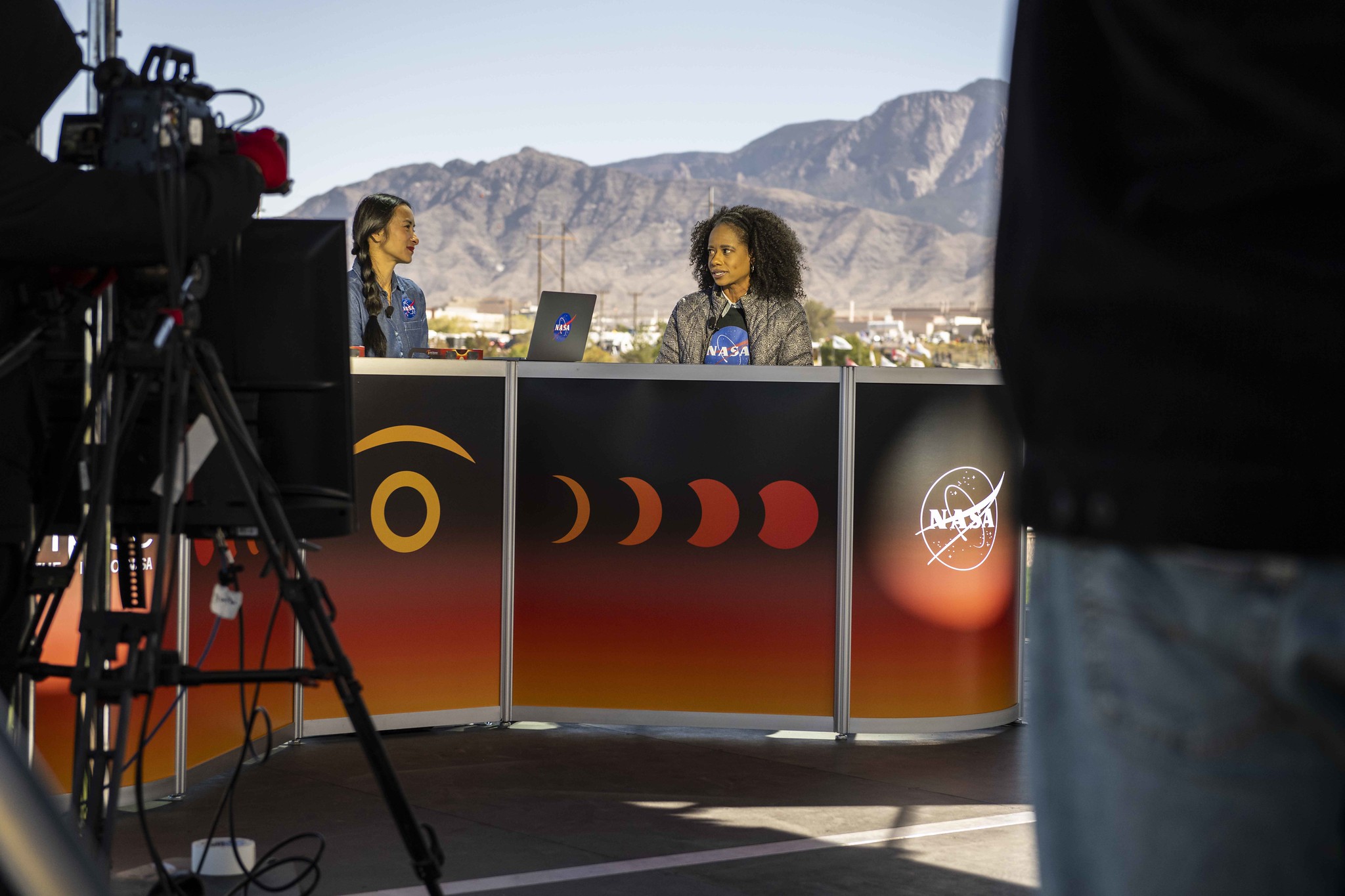 Two women sit at a desk outside. The desk is decorated with art that represents an annular eclipse. There are people and a camera recording them. In the background are mountains.