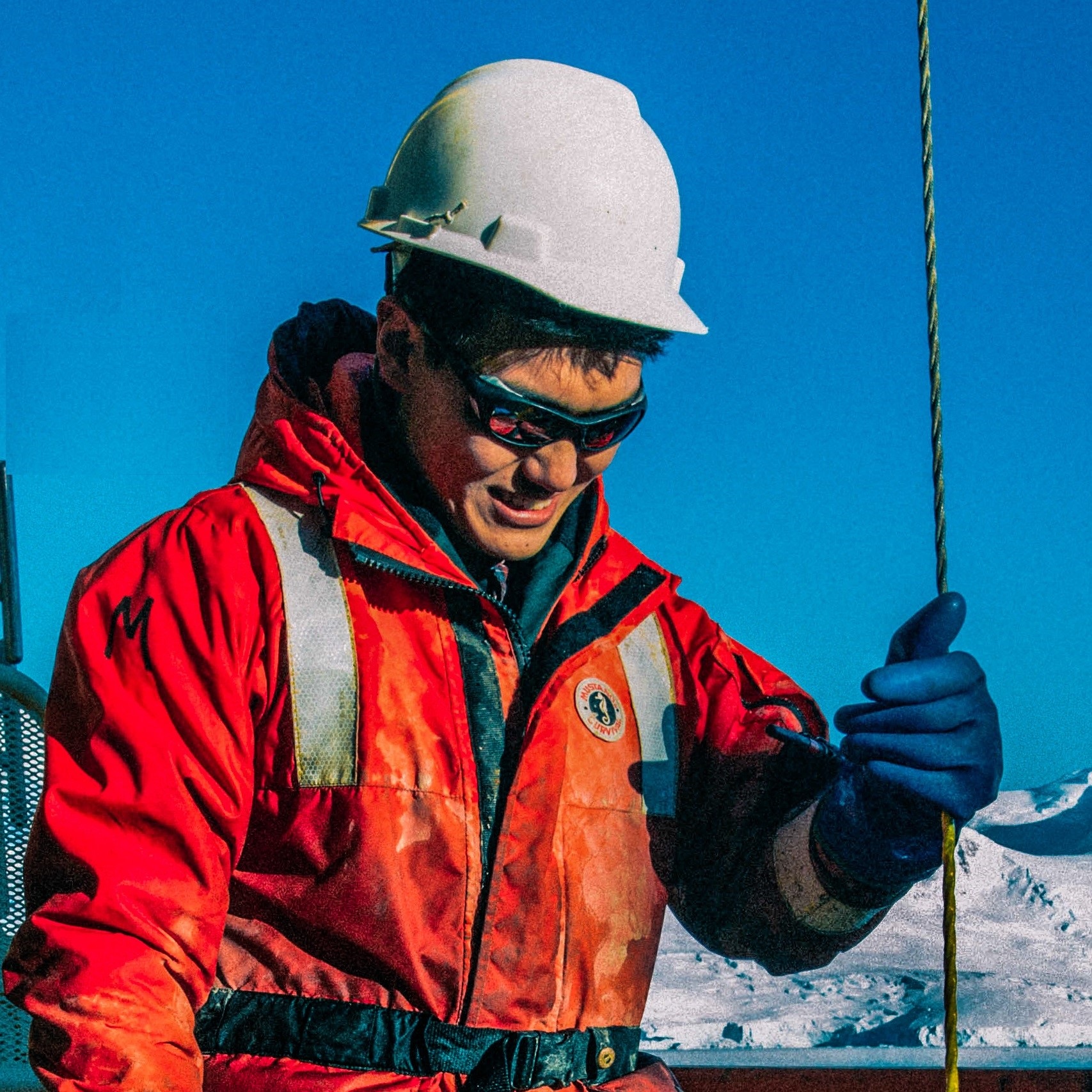 Photo of a man in an orange winter jacket and a white hard hat holding a rod in his left hand.