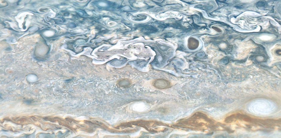 Big swirls, little swirls, spots, and streams of clouds swirl across this image of the gaseous atmosphere of Jupiter. The top of image is mostly blues and whites, the middle third has smaller, tighter curls and occasional spots in tans, whites, and blues. The bottom edge is dominated by a brownish, snaky line of clouds.