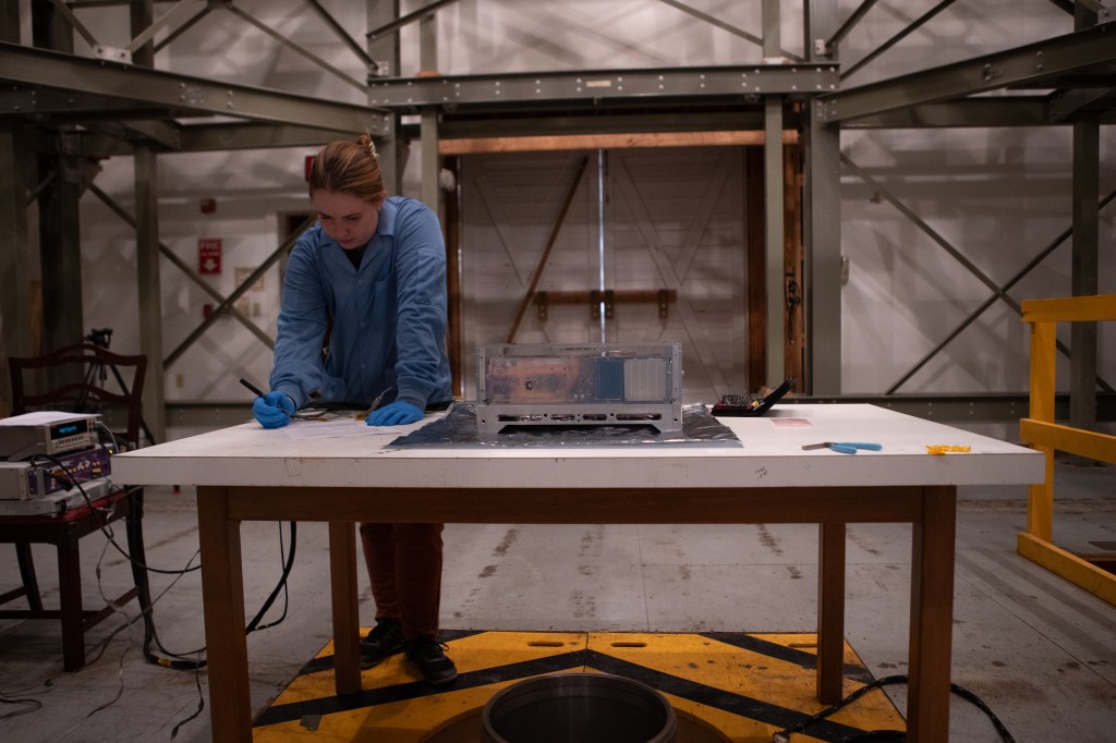 An engineer fills out a form on a table hosting a small shiny spacecraft.