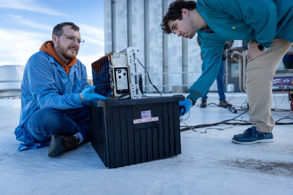 This photograph shows two men on a rooftop working on a small spacecraft.