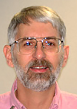 Portrait photo of a man with grey hair, a beard and glasses wearing a pink shirt.
