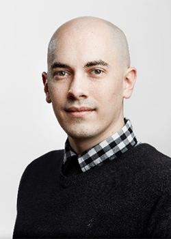 Portrait photo of a bald man with a slight smile wearing a black and white checkered collared shirt under a black sweater.