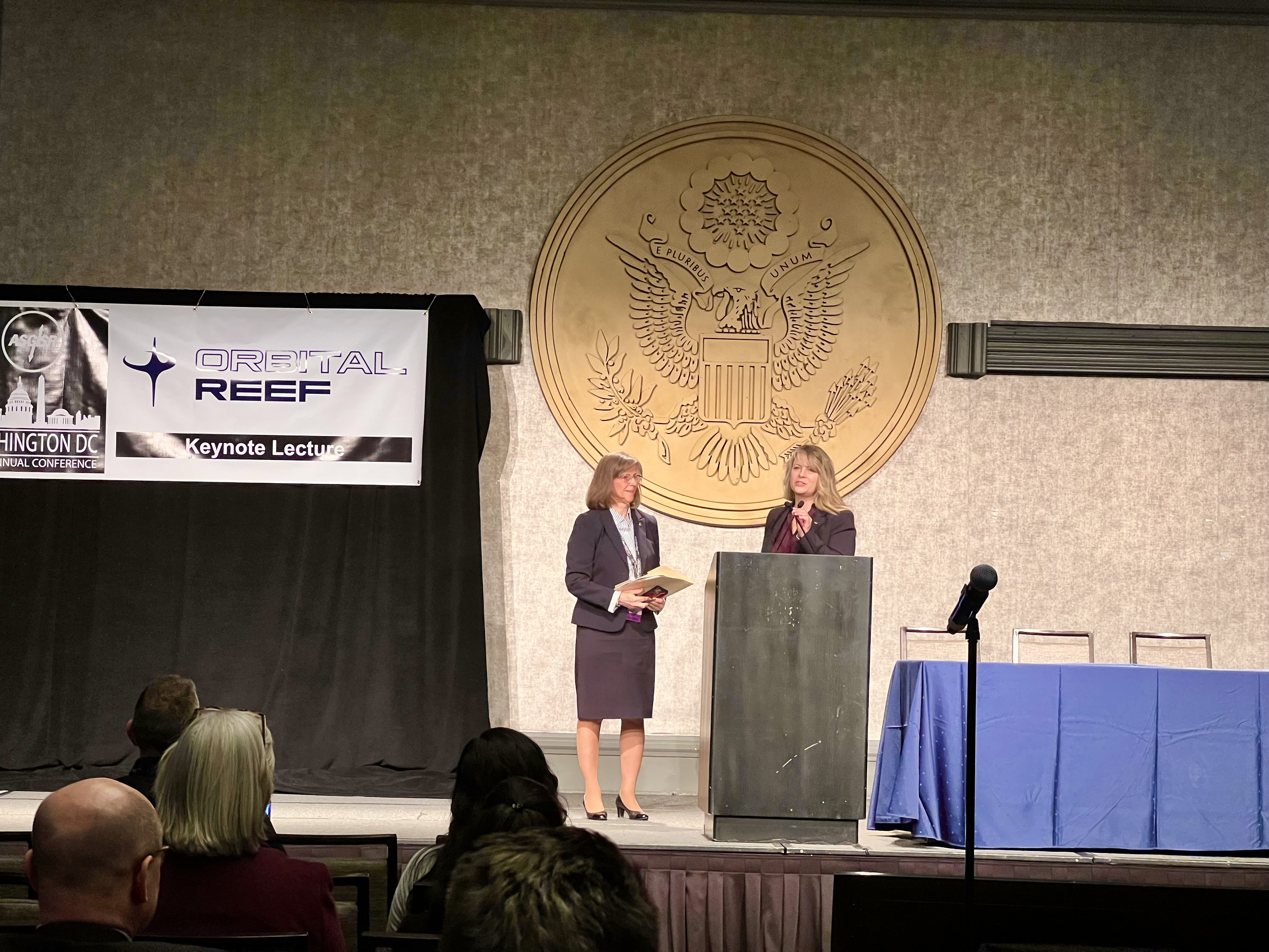 Dr. Lisa Carnell (right) and Dr. Bonnie Dunbar (left) on stage presenting at the main conference podium. Both women are wearing dark, formal business attire.